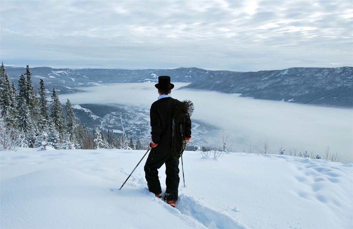 Feiaren på veg til hytta i snø, ser utover fjell og delvis tåkelagt tettstad - Klikk for stort bilete